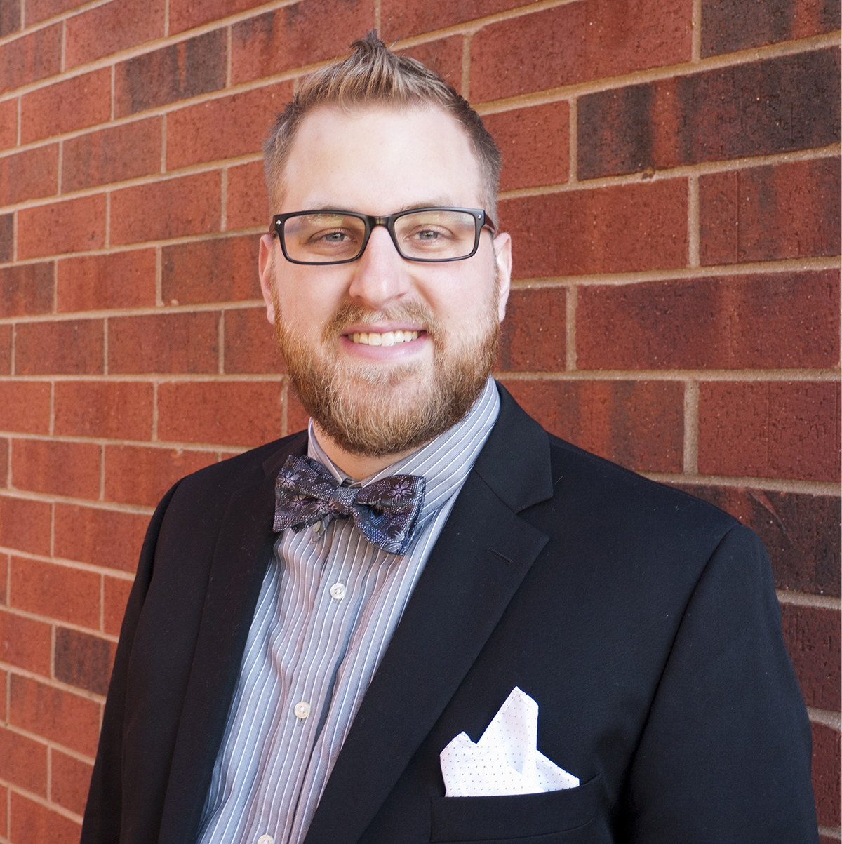 a headshot of Chad O'Brien, a young white man with a beard, wearing glasses, and dressed in a suit and bowtie.
